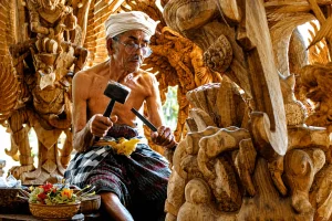An elderly Balinese artisan carefully creating traditional Balinese wood carving by hand, surrounded by intricate wooden sculptures and detailed cultural patterns in a rustic workshop in Bali.