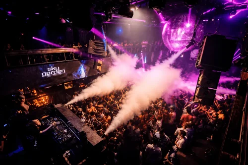 Crowded nightclub party at Sky Garden in Kuta Bali with DJ, colorful lights, and smoke effects on the dance floor.