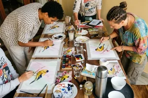 People painting colorful birds on canvas at a wooden table, showcasing the creative variety found in Bali art workshops | balitouristic.com
