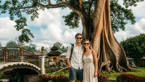 A smiling British couple posing in front of a large Bayan ancient tree in a lush Balinese garden, with a stone bridge and temple structures under a partly cloudy sky.