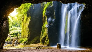 A stunning view of Goa Raja Waterfall featuring powerful cascading water flowing through lush jungle cliffs into a clear natural pool surrounded by tropical greenery and misty scenery.