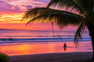 A stunning sunset view at Kelating Beach featuring glowing orange skies, reflective black sand, dramatic ocean waves, and a peaceful tropical coastline at dusk.