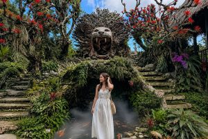A female tourist in a white dress posing in front of a giant, intricate lion head wood carving over a misty pond at sari timbul glass factory. | www.balitouristic.com