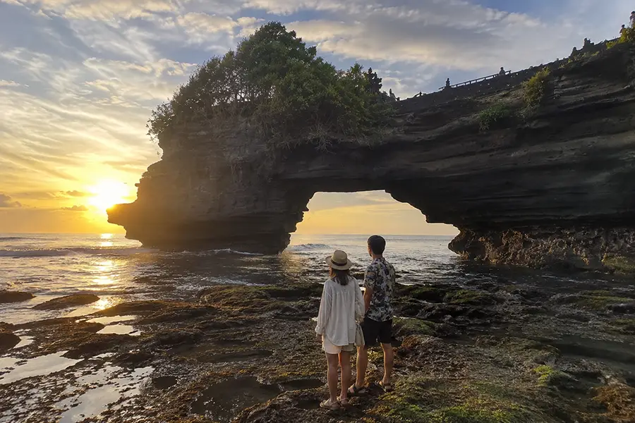 A romantic couple standing together on the dark stone, admiring a breathtaking golden sunset over the ocean horizon at batu bolong beach.