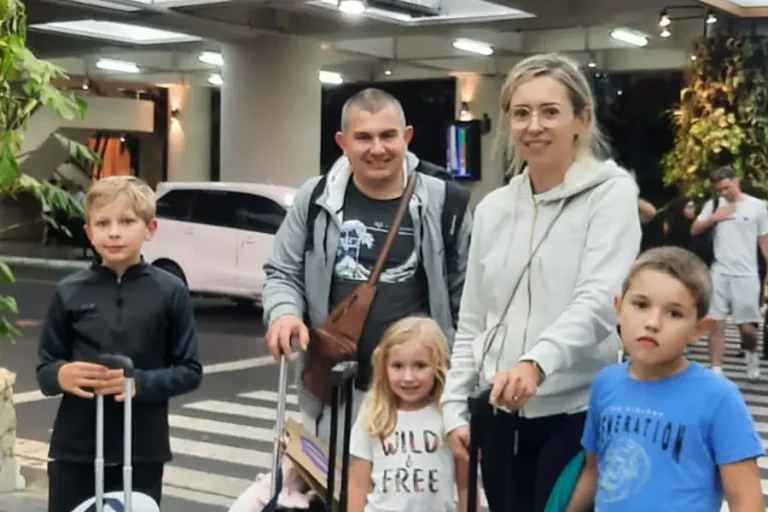 A family with luggage at the airport pickup lane at night, highlighting the best bali airport transfer service for late arrivals | balitouristic.com