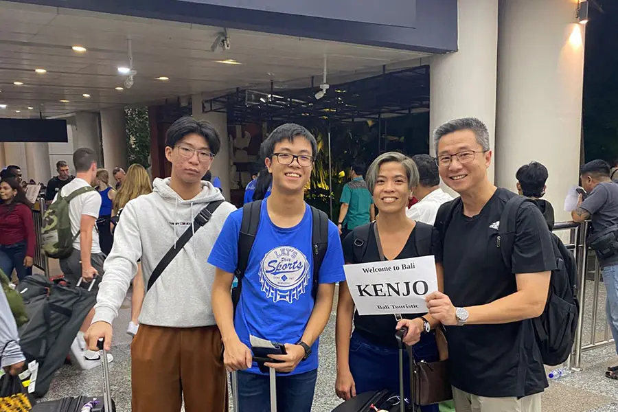 A group of tourists holding a Bali Touristic name sign during a late-night arrival, showcasing the best bali airport transfer service | balitouristic.com