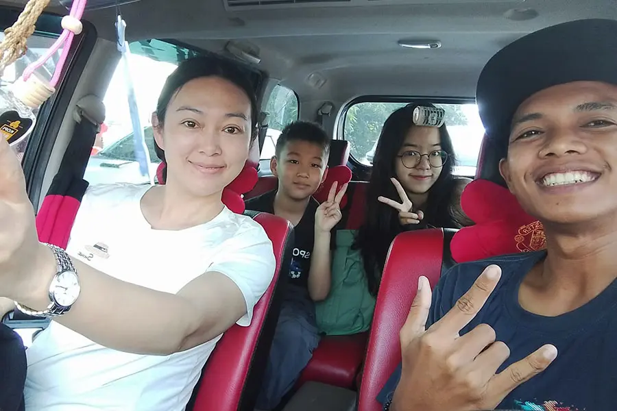 A happy family taking a cheerful selfie with their professional local chauffeur inside a comfortable private car while choosing to hire driver bali. | balitouristic.com