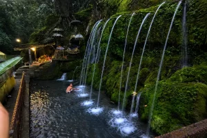 A tourist participating in the traditional Melukat purification ritual, standing under a natural water spout at tirta sudamala temple. | balitouristic.com