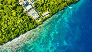A stunning bird’s-eye view of Toya Pakeh Beach showcasing crystal-clear turquoise waters, vibrant coral reef areas, traditional boats, and beautiful tropical coastline scenery from above.