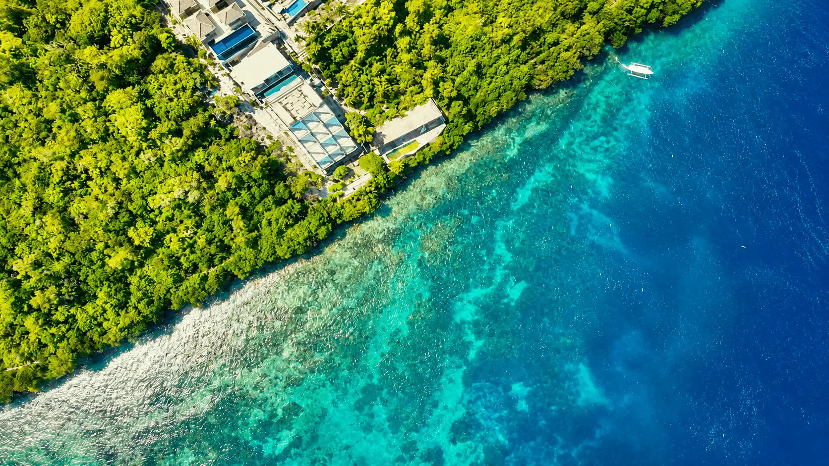 A stunning bird’s-eye view of Toya Pakeh Beach showcasing crystal-clear turquoise waters, vibrant coral reef areas, traditional boats, and beautiful tropical coastline scenery from above.