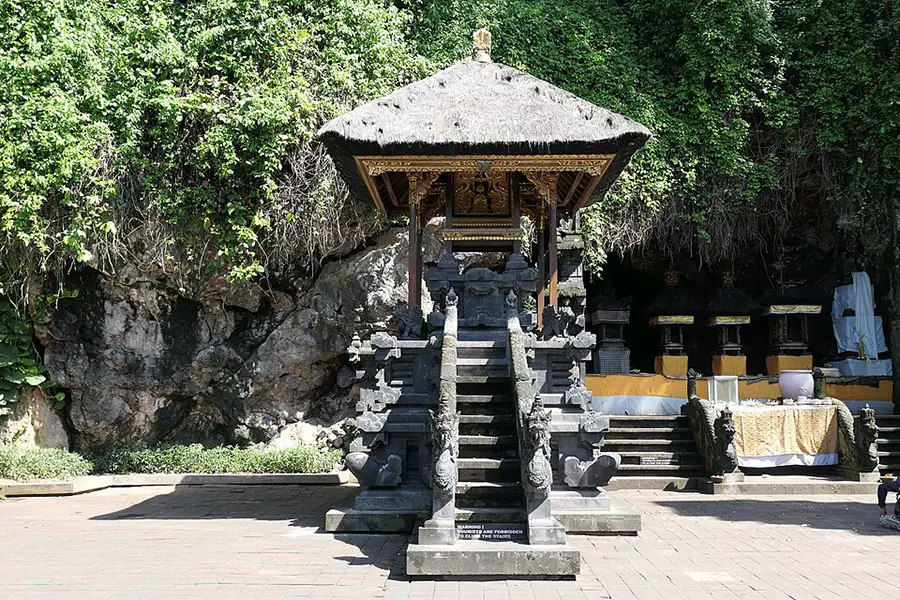 A multi-tiered stone shrine with a thatched roof situated directly in front of a dark rocky cave cliff at goa lawah temple.