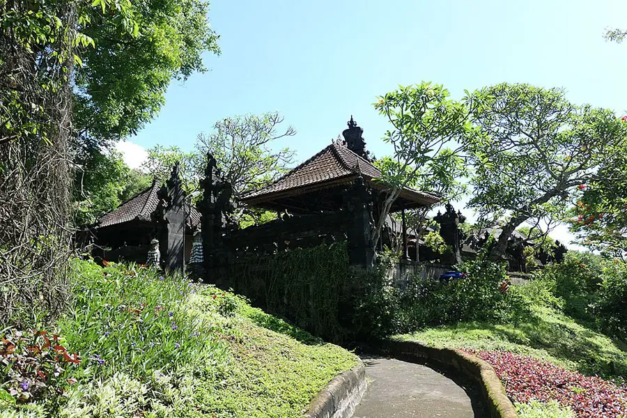 An outdoor pathway winding past tiered black stone pavilions and lush blooming gardens at goa lawah temple.