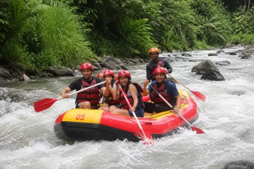 Thrilled tourists wearing red helmets and life jackets paddling a red and yellow inflatable raft through river rapids, showing a fun water sport option on an all-inclusive bali trip.