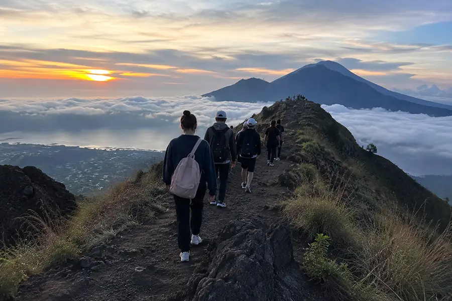 A group of travelers with backpacks hiking along a narrow mountain ridge at dawn during an active sunrise trek, a popular adventure activity included in an all-inclusive bali vacation package. | balitouristic.com