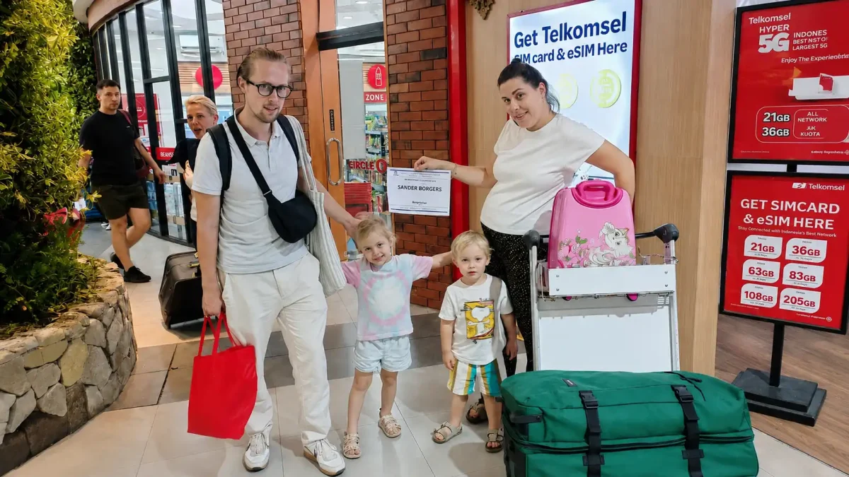 A smiling European family being warmly welcomed by a friendly local driver during an airport pickup in Bali, standing beside a spacious modern MPV with luggage and ready to begin their tropical island vacation.