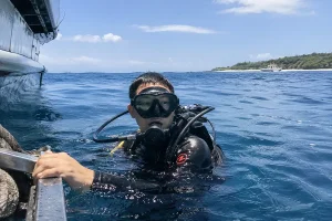 A male scuba diver wearing full gear and a mask holding onto a boat ladder in the open blue ocean water, capturing an exciting excursion with bali jet set dive.