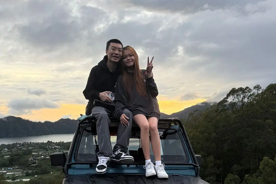 A happy couple sitting on the hood of a 4x4 tour jeep while enjoying a golden sunrise over the lake and mountains, a scenic highlight of bali packages for couple.