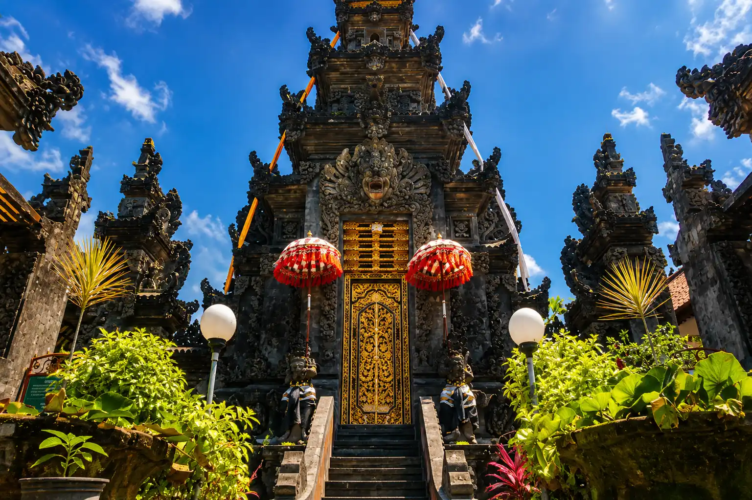 A peaceful view of Blanjong Temple featuring traditional Balinese architecture, ancient stone carvings, lush tropical gardens, and a serene spiritual atmosphere surrounding the historic Blanjong Pillar.