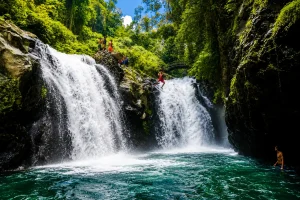 An adventurous traveler cliff jumping into the natural pool at Aling-Aling Waterfall, surrounded by cascading waterfalls, lush tropical rainforest, dramatic rock formations, and an exciting outdoor adventure atmosphere in North Bali.