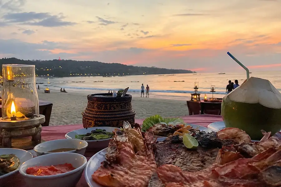 A close-up of a fresh seafood platter with grilled prawns, fish, a whole coconut, and a candle lantern on a table during dinner at jimbaran beach.