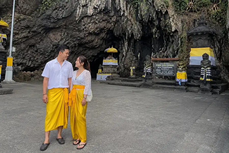 A tourist couple wearing traditional yellow ceremonial sashes and sarongs in front of the sacred bat cave at Pura Goa Lawah during an east bali tour. | balitouristic.com