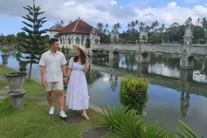 A traveler couple holding hands and walking along the grassy lakeside paths of Taman Ujung Water Palace during an east bali tour. | balitouristic.com
