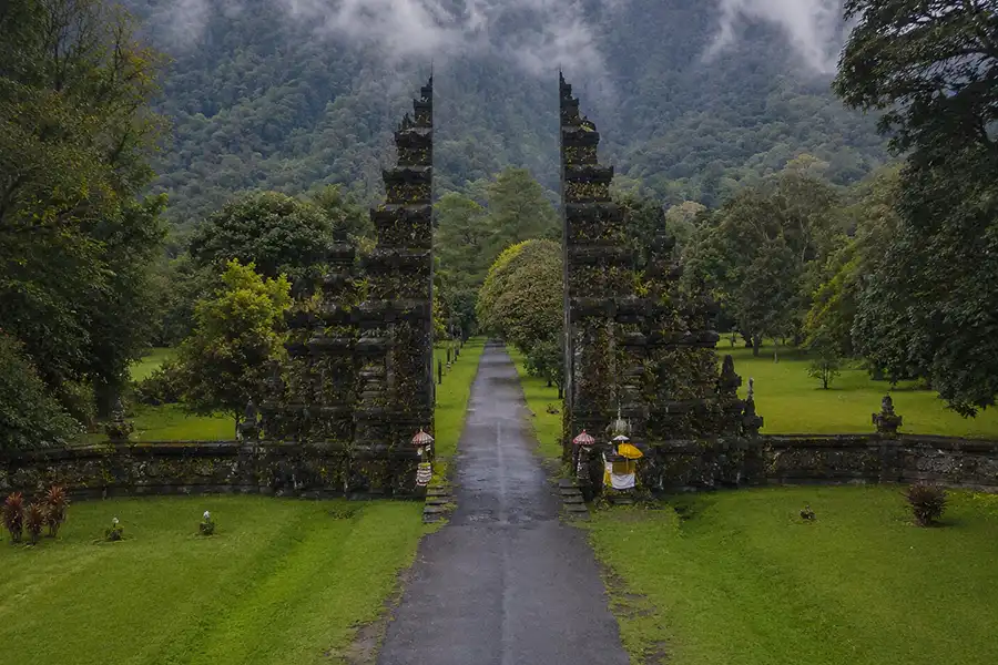 An aerial view of a traditional stone split gateway opening up to a long paved pathway surrounded by green lawns and misty mountains, one of the most famous bali tourist spots.