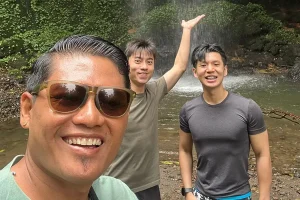 friendly local driver taking a smiling group selfie with two male tourists at a hidden jungle waterfall after they decided to hire bali driver ubud. | balitouristic.com