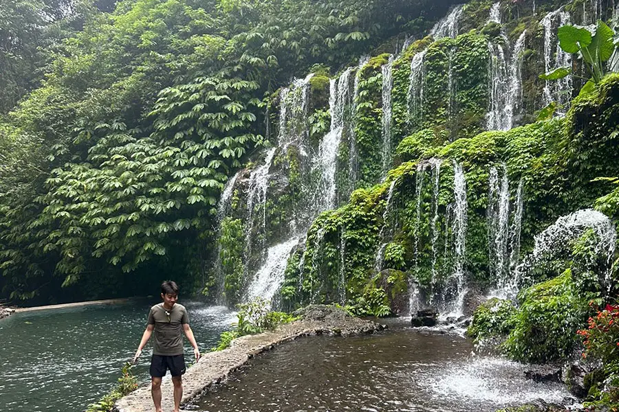 A tourist standing on a stone pathway next to a magnificent, multi-tiered jungle waterfall, illustrating a popular route when you hire bali driver ubud. | balitouristic.com