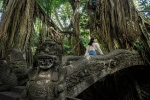 A female traveler walking across an ancient stone dragon bridge surrounded by massive banyan tree roots at a famous Interest Place Bali. | balitouristic.com