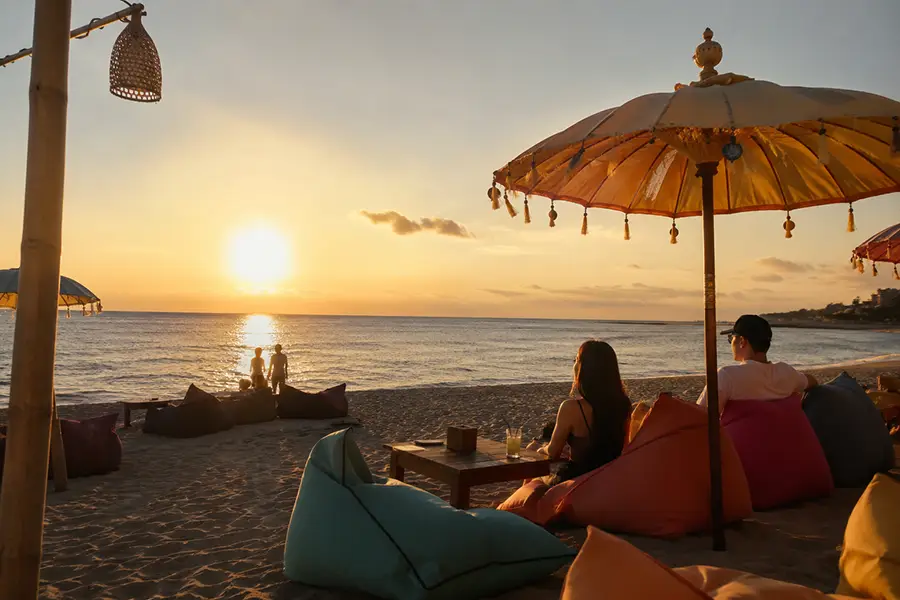 Tourists relaxing on colorful bean bags under traditional umbrellas on the beach while watching a golden sunset, showcasing a popular Interest Place Bali. | balitouristic.com