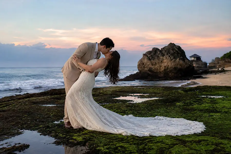 A romantic couple sharing a dip kiss on a mossy coastal rock formation during sunset near the ocean, showcasing beautiful and interesting places in bali. | balitouristic.com