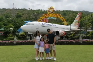 A happy family of four posing for a holiday photo on a green lawn in front of the iconic Krisna Air plane at krisna funtastic land. | balitouristic.com