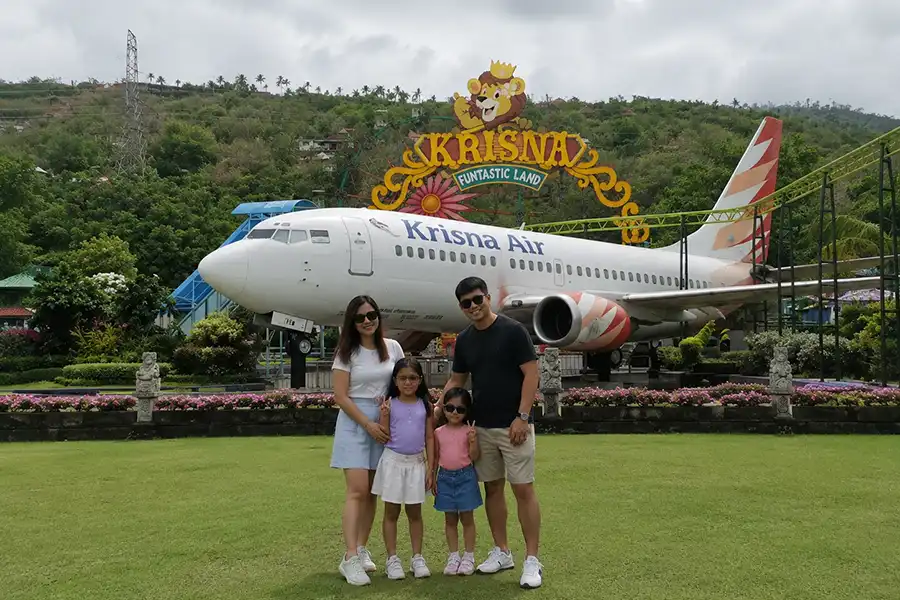 A happy family of four posing for a holiday photo on a green lawn in front of the iconic Krisna Air plane at krisna funtastic land. | balitouristic.com