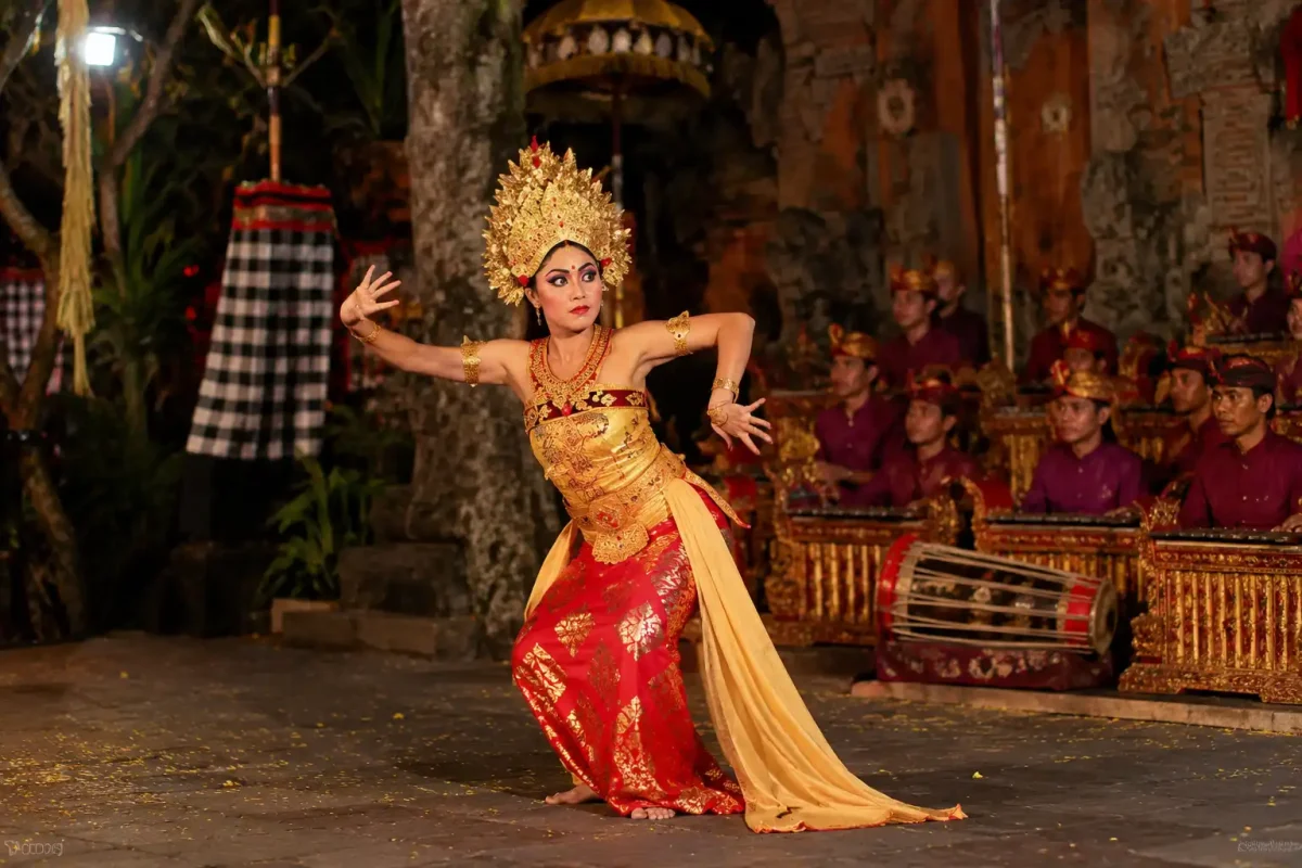 A traditional Balinese female dancer in a striking red and gold costume performing the graceful legong dance bali with a live gamelan orchestra in the background.