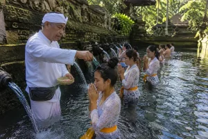 A traditional Melukat ceremony in Bali featuring a Balinese pemangku (temple priest) guiding participants through sacred cleansing rituals with holy water, traditional offerings, temple shrines, and a serene spiritual atmosphere surrounded by lush tropical surroundings.