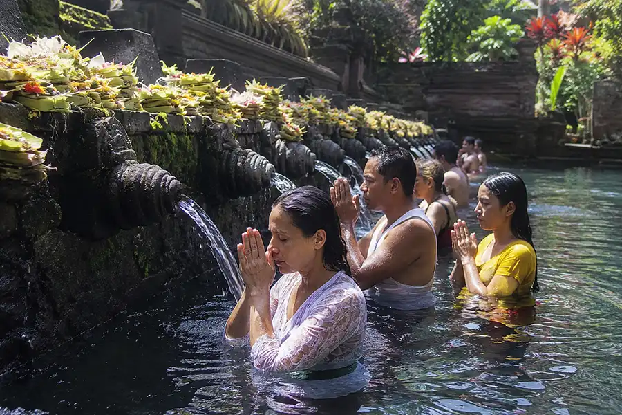 A group of visitors participating in a traditional Melukat ceremony in Bali at Tirta Empul Temple, receiving blessings and cleansing under the sacred holy water fountains, surrounded by ancient temple architecture, ceremonial offerings, and a serene spiritual atmosphere.