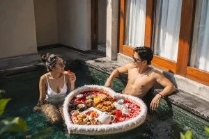 A smiling young couple enjoying a luxurious floating breakfast served in a heart-shaped basket inside their private villa plunge pool during a package bali honeymoon. | balitouristic.com
