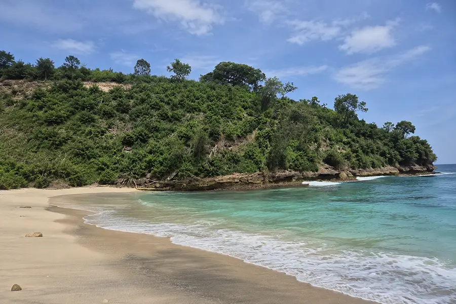 A pristine white sand shore meeting clear turquoise waves beneath a steep, lush green hill at pandan beach bali.