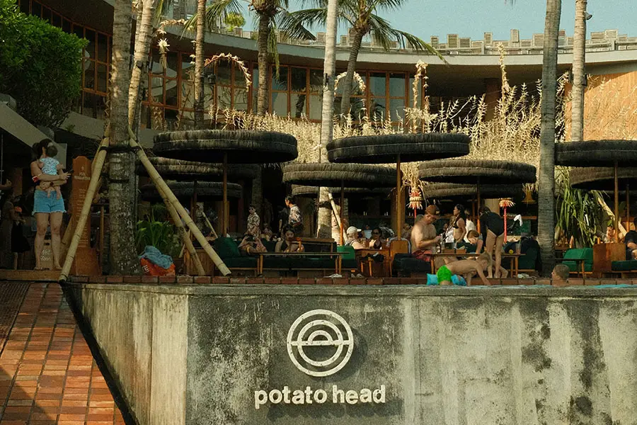 Guests relaxing around the swimming pool area behind a rustic concrete wall displaying the circular logo of potato head bali.