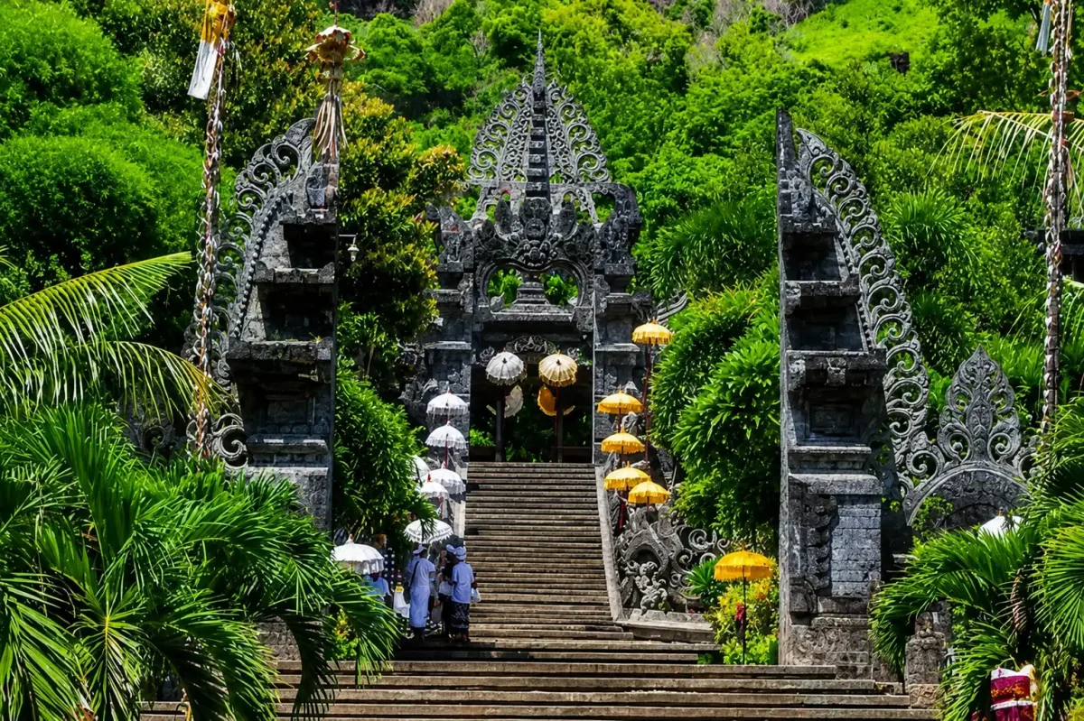 Balinese worshippers standing at the bottom of a grand stone staircase leading up to the ornate split gates of the sacred pulaki temple. | balitouristic.com