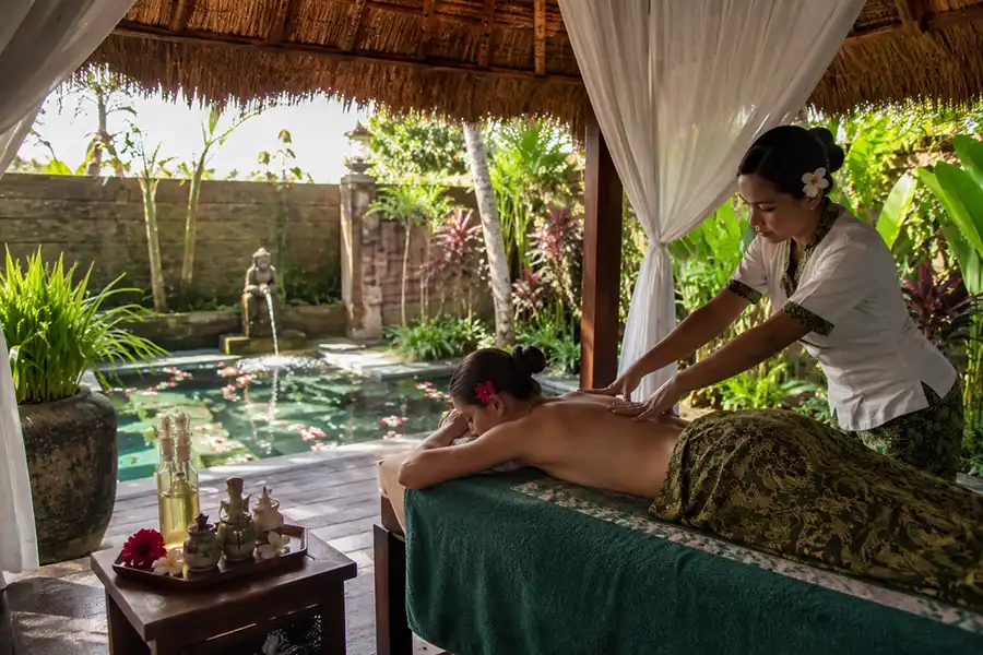 A female tourist enjoying a professional back massage from a local spa therapist in an outdoor pavilion overlooking a tropical garden pool, a classic element of a relaxing vacation in bali. | balitouristic.com