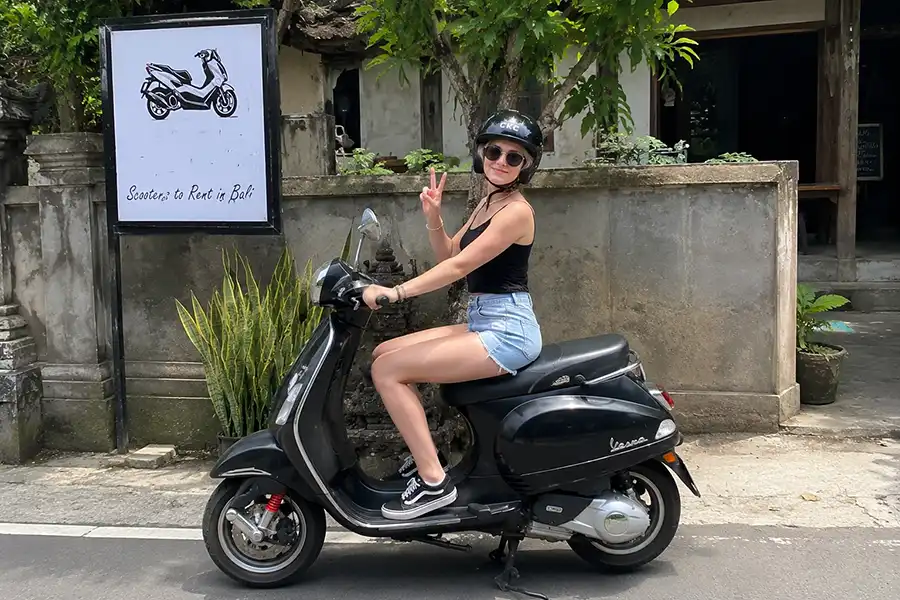 A female tourist sitting on a classic black Vespa scooter and giving a peace sign next to a rental sign for scooters in bali. | balitouristic.com