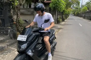A man in a helmet sitting on a parked black Yamaha NMAX maxi-scooter along a quiet village street, showcasing premium models for scooters in bali. | balitouristic.com