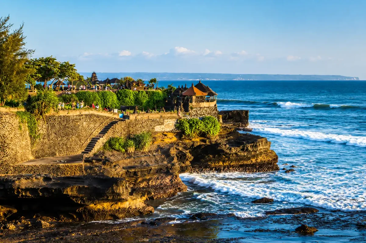 A dramatic view of Tanah Lot Temple during a Tanah Lot Sunset Tour, featuring the iconic sea temple rising from rugged coastal rocks, powerful ocean waves crashing below, vibrant golden-orange sunset skies, and breathtaking panoramic scenery that captures the magic of Bali’s coastline.