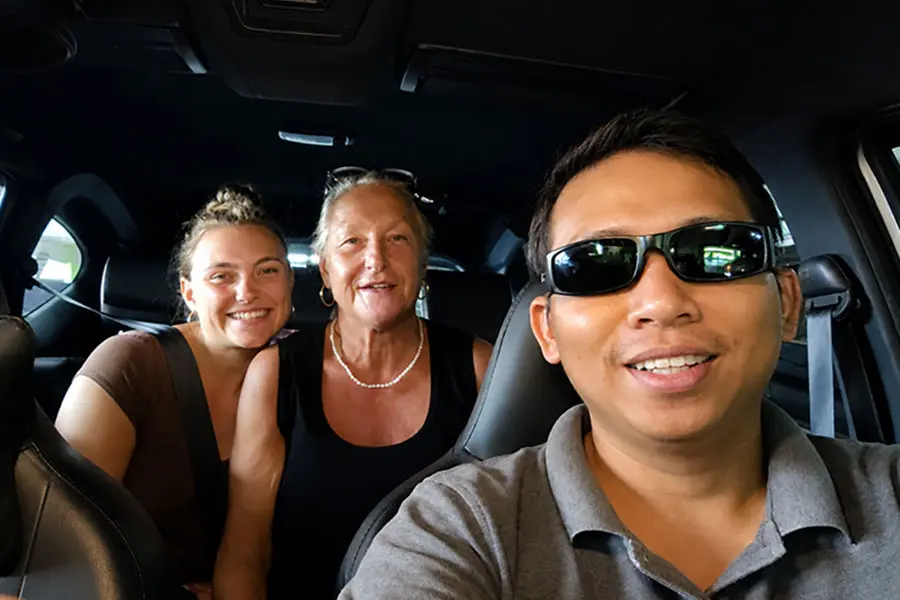 A professional local driver smiling alongside two happy international tourists inside a clean, modern private car for an elite taxi airport bali alternative. | balitouristic.com