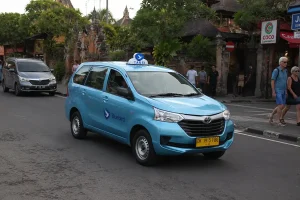 A distinctive light blue Bluebird taxi driving down a main street past pedestrians and traditional Balinese buildings, showing a popular taxi airport bali choice. | balitouristic.com