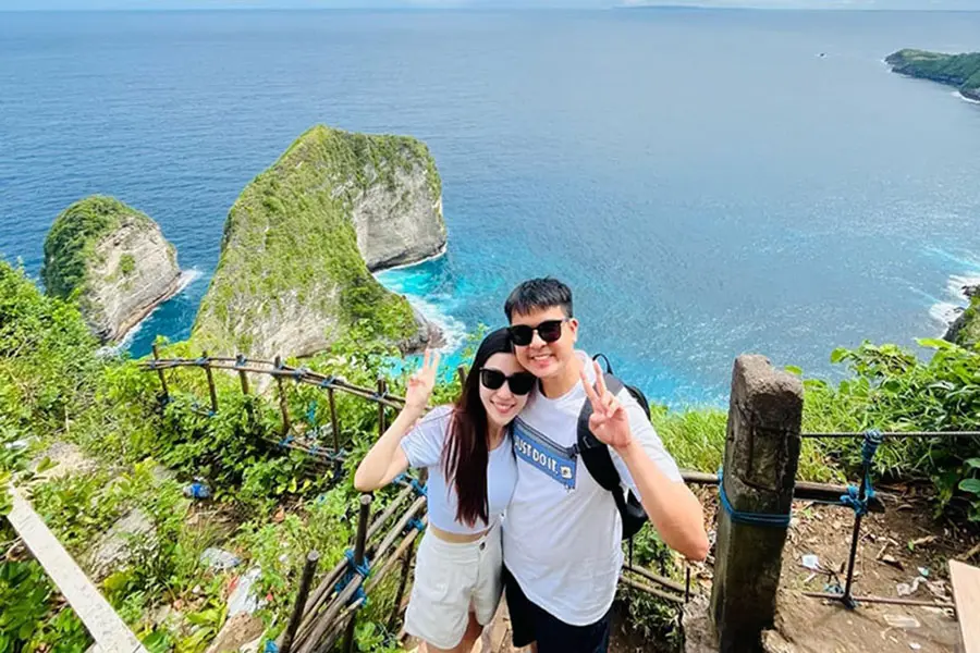 A happy couple standing on a cliffside viewpoint looking down at the famous dinosaur-shaped Kelingking Beach cliff, a must-visit idea for things to do in bali for couples. | balitouristic.com