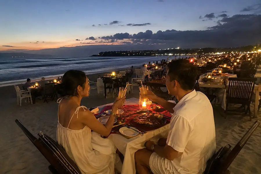 A couple enjoying a private candlelit seafood dinner at a wooden table on the beach during a golden sunset, a perfect choice for things to do in bali for couples. | balitouristic.com