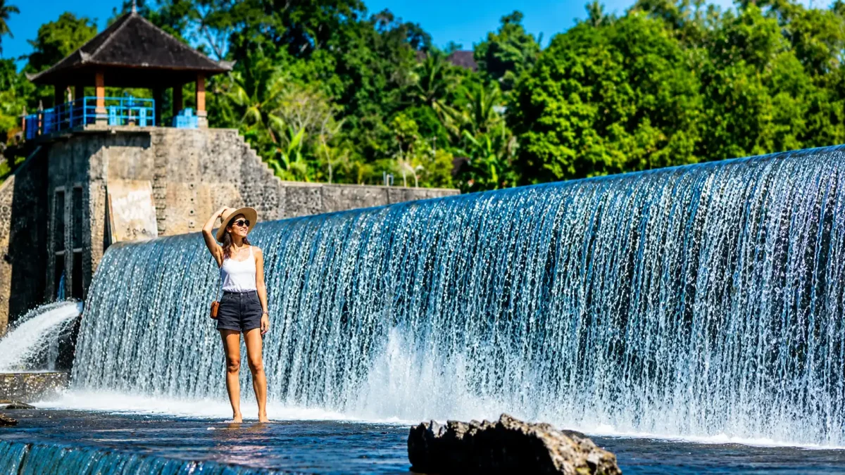 tukad unda - klungkung interesting places to visit - bali touristic Visitors relax and take photos at Tukad Unda Waterfall, where wide cascading water flows over a stone dam surrounded by lush greenery. The image showcases one of Bali’s unique waterfalls, offering a peaceful atmosphere and picturesque scenery for travelers.